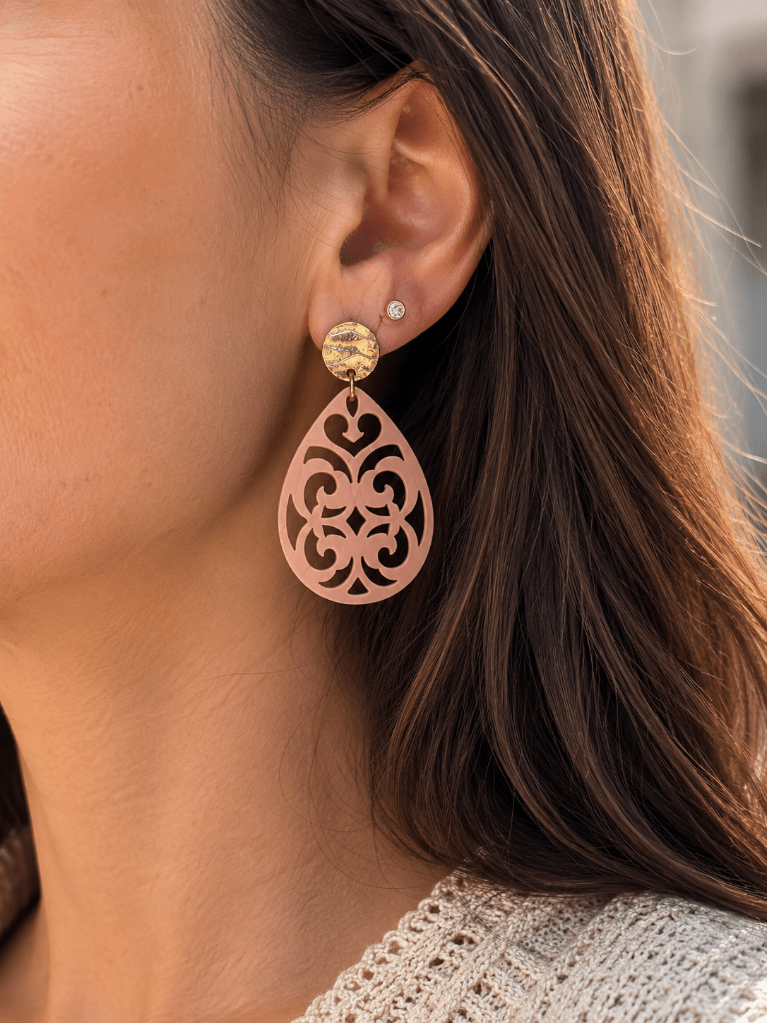 Boucles d'oreilles pendantes en acrylique rose poudré découpé à motif arabesque, avec puce ronde dorée martelée, portées par une femme aux cheveux châtains