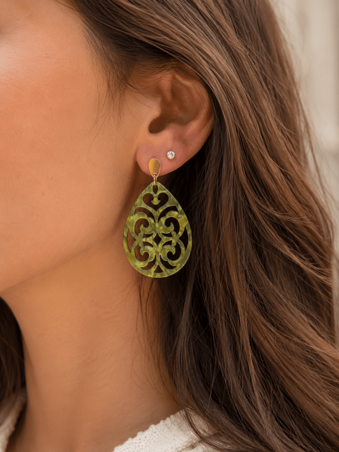 Boucles d'oreilles pendantes en acrylique vert marbré découpé à motif arabesque, avec attache dorée ovale, portées par une femme aux cheveux bruns