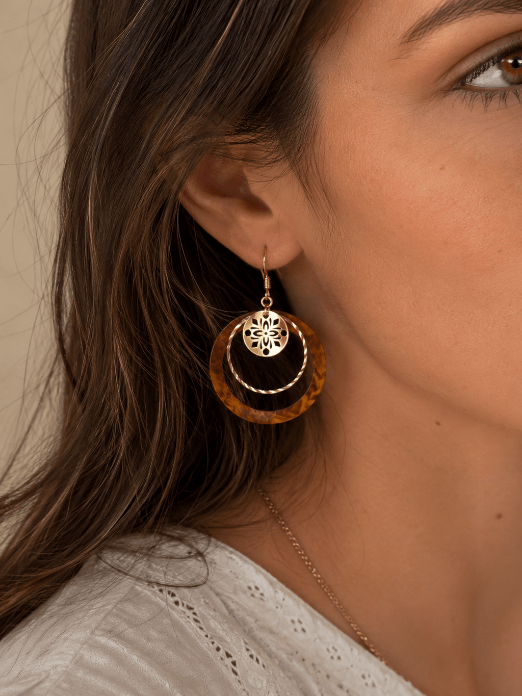 Boucles d'oreilles pendantes composées d'un cercle acrylique effet écaille caramel et d'un anneau doré martelé, avec breloque ronde ajourée dorée, portées par une femme