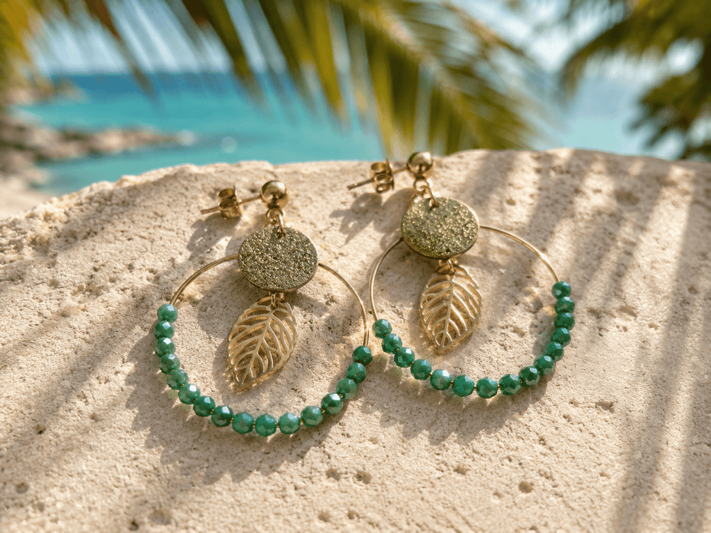 Boucles d'oreilles créoles dorées avec médaillon pailleté, breloque feuille ajourée et perles facettées vertes, posées sur sable blanc avec mer turquoise en arrière-plan