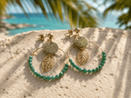 Boucles d'oreilles créoles dorées avec médaillon pailleté, breloque feuille ajourée et perles facettées vertes, posées sur sable blanc avec mer turquoise en arrière-plan