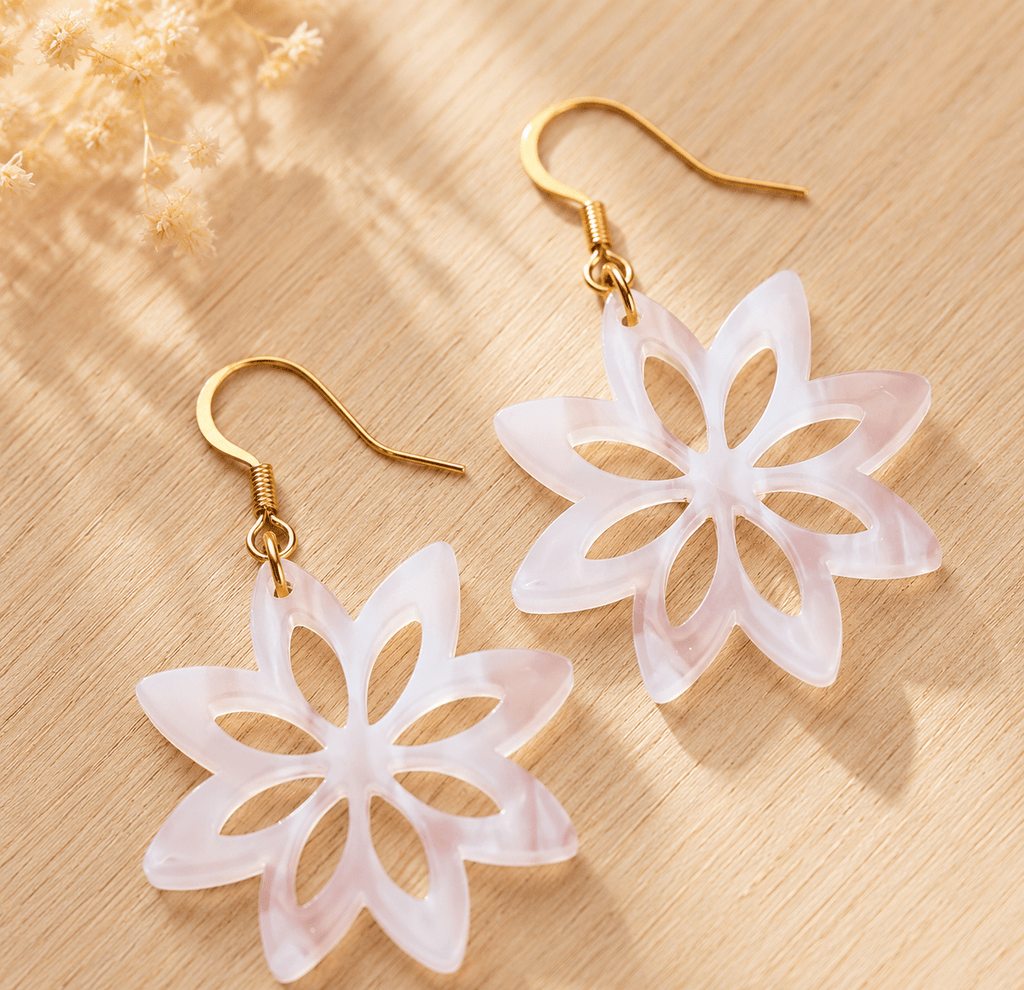 Paire de boucles d'oreilles fleur en acrylique nacré rosé, pétales découpés, crochets dorés, posées sur bois clair avec fleurs séchées