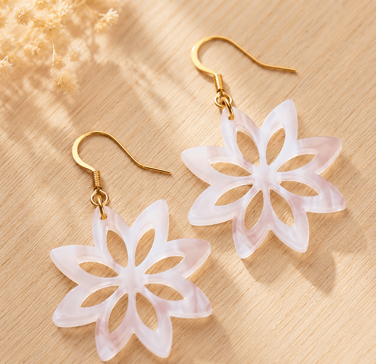 Paire de boucles d'oreilles fleur en acrylique nacré rosé, pétales découpés, crochets dorés, posées sur bois clair avec fleurs séchées