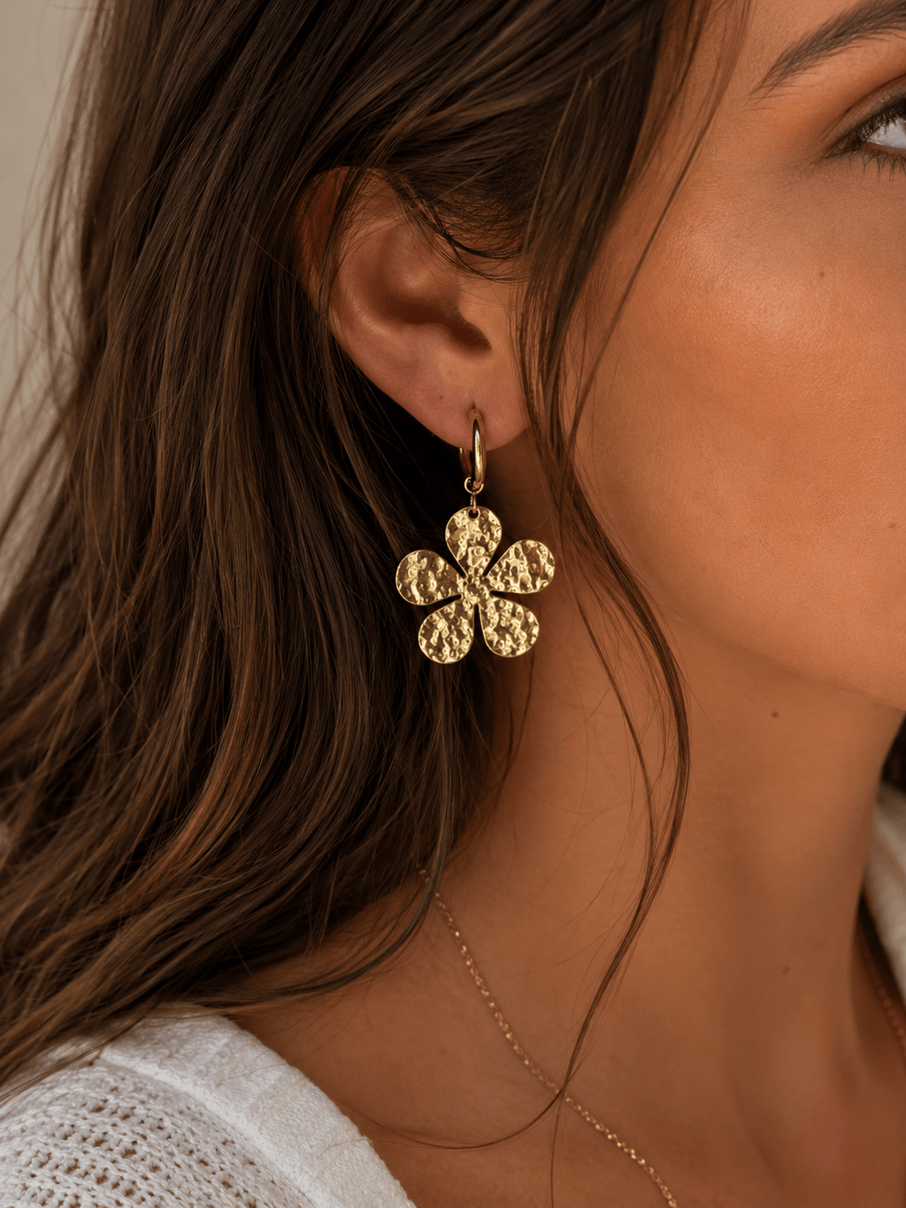 Boucles d'oreilles créoles dorées avec charm fleur martelée à 6 pétales ronds, portées par une femme aux cheveux ondulés