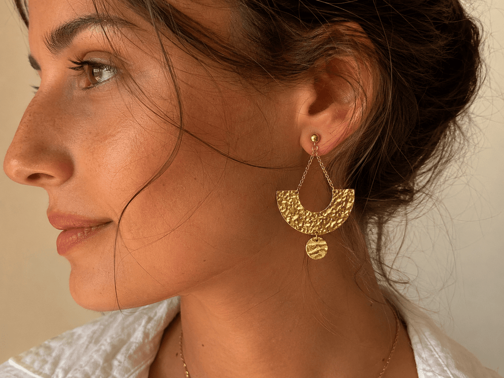 Boucles d'oreilles dorées martelées en forme de croissant, suspendues par une chaîne fine à une puce boule dorée, portées par une femme aux cheveux relevés