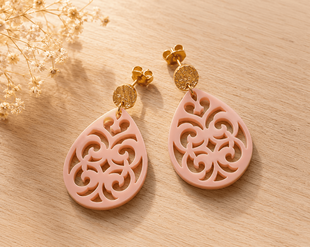 Paire de boucles d'oreilles en acrylique rose nude en forme de goutte ajourée motif baroque, montées sur puces dorées texturées, fond bois clair
