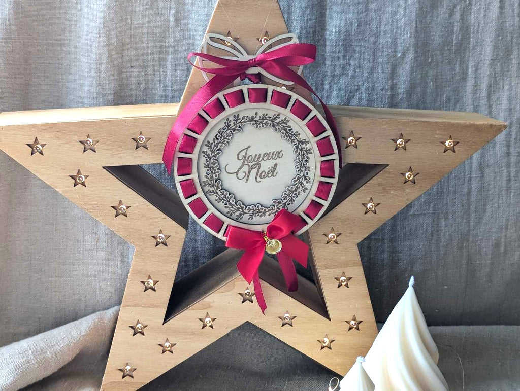 Boule de Noël personnalisée en bois naturel gravé avec ruban rouge profond entrelacé effet cocarde, texte Joyeux Noël et couronne de feuillage gravée au laser