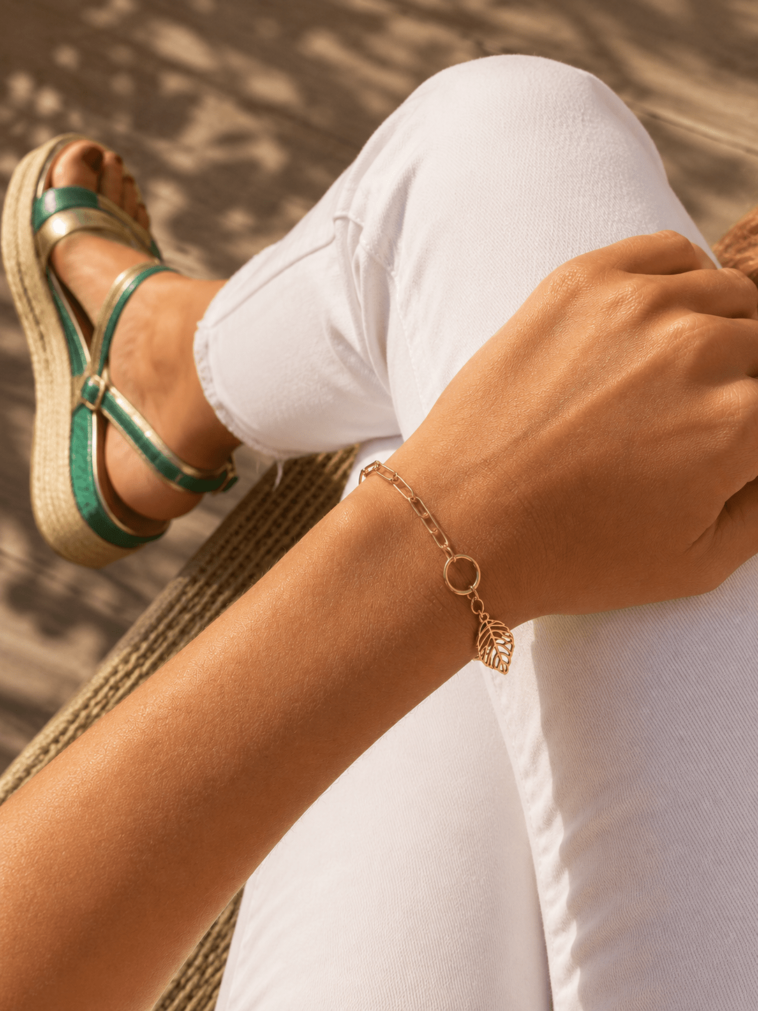 Bracelet chaîne dorée à maillons avec anneau et breloque feuille ajourée, porté au poignet d'une femme en pantalon blanc, ambiance estivale