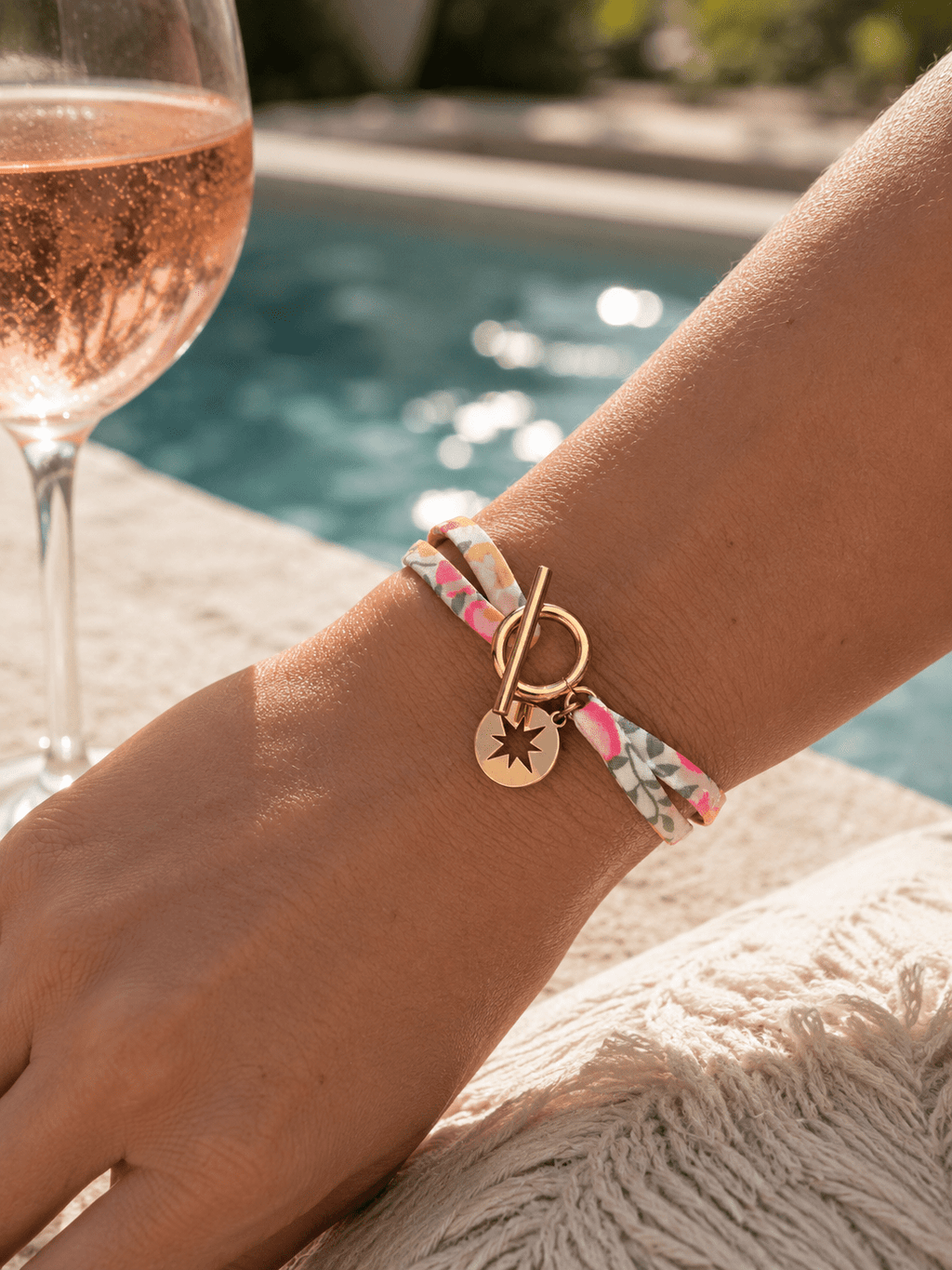 Bracelet liberty fleuri rose et vert double tour avec fermoir toggle doré et breloque médaillon étoile, porté au poignet au bord d'une piscine ensoleillée