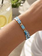 Gros plan sur bracelet liberty turquoise à motif floral avec perle tube et anneaux dorés, porté au poignet, verre de limonade en arrière-plan