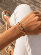 Stack de deux bracelets dorés au poignet : bracelet chaîne avec breloque feuille ajourée et bracelet perles vertes avec trèfle doré, ambiance été