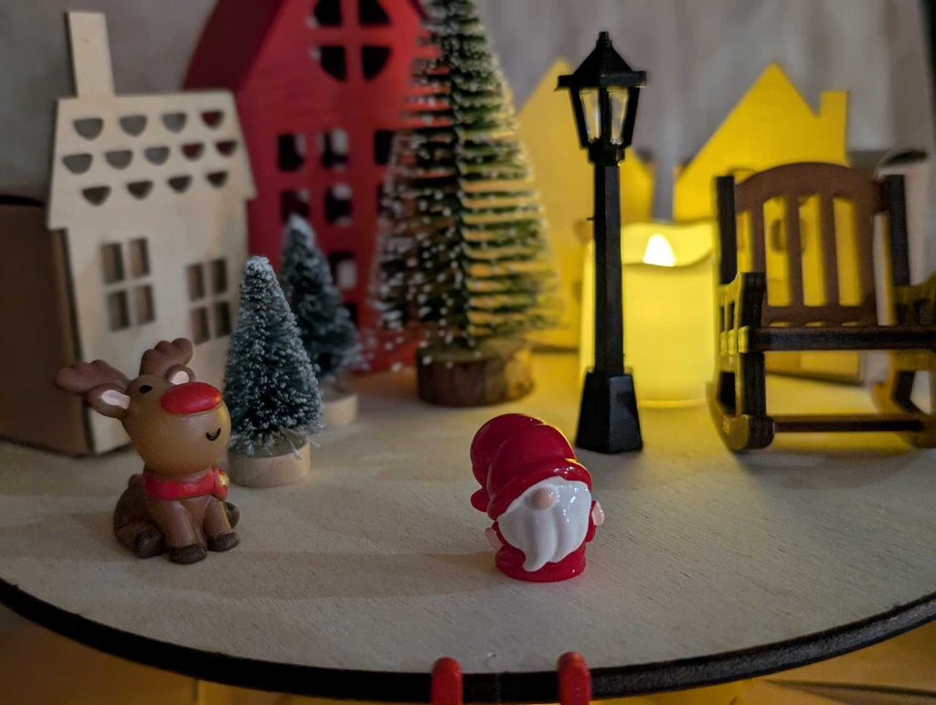 Détail décorations calendrier de l'avent : figurine renne kawaii et Père Noël rouge, mini sapin enneigé, lampadaire noir miniature et maisonnettes illuminées en arrière-plan