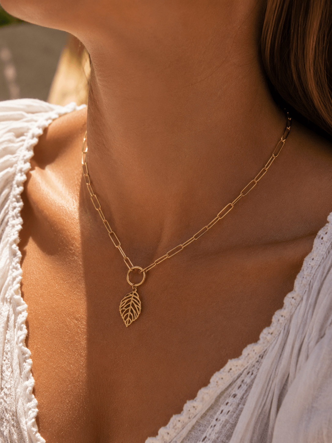 Collier chaîne dorée à grands maillons avec anneau et pendentif feuille ajourée dorée, porté au décolleté d'une femme en top blanc brodé, lumière solaire