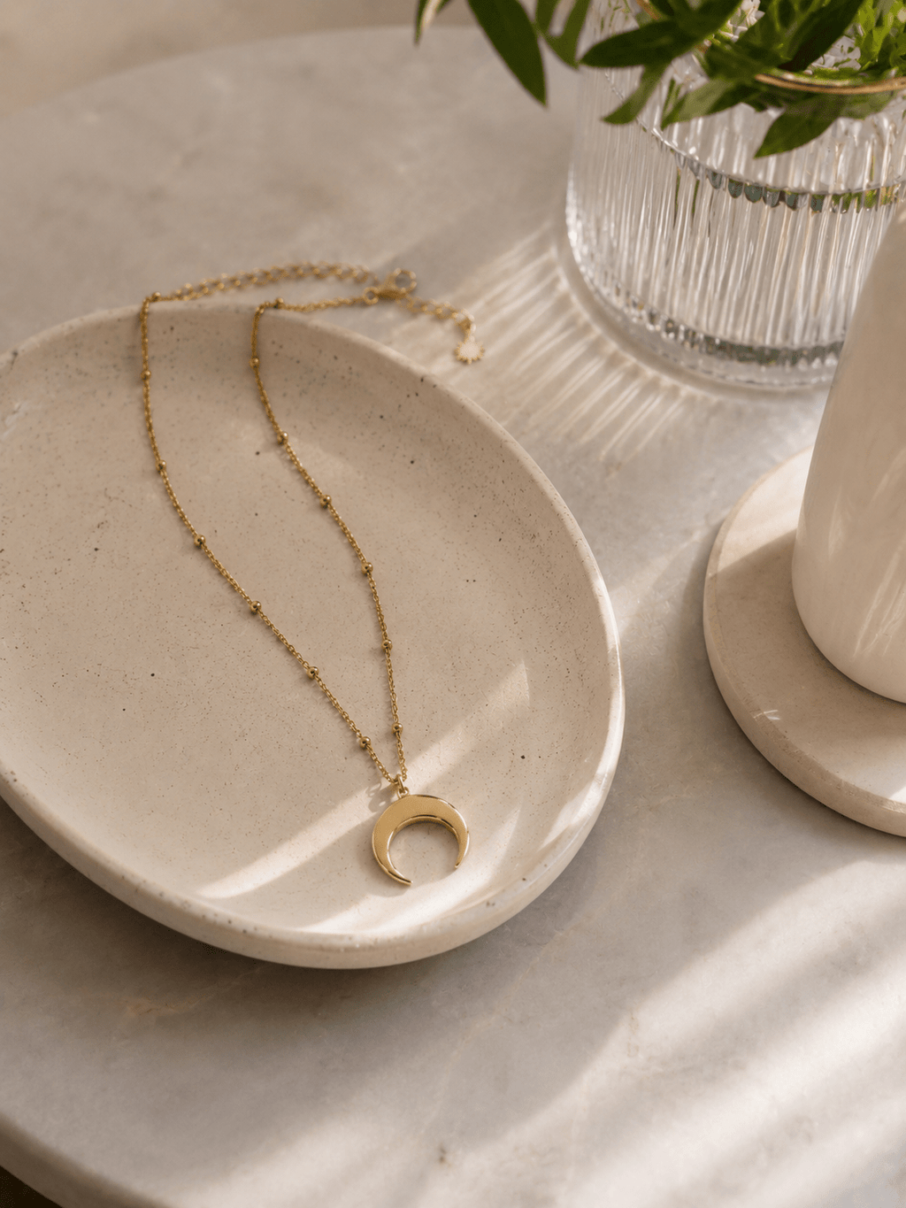 Collier corne lune doré posé dans une coupelle en pierre sur marbre, bijou lunaire acier doré ambiance épurée