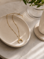 Collier corne lune doré posé dans une coupelle en pierre sur marbre, bijou lunaire acier doré ambiance épurée
