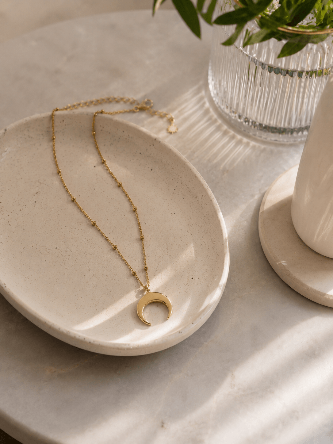 Collier corne lune doré posé dans une coupelle en pierre sur marbre, bijou lunaire acier doré ambiance épurée