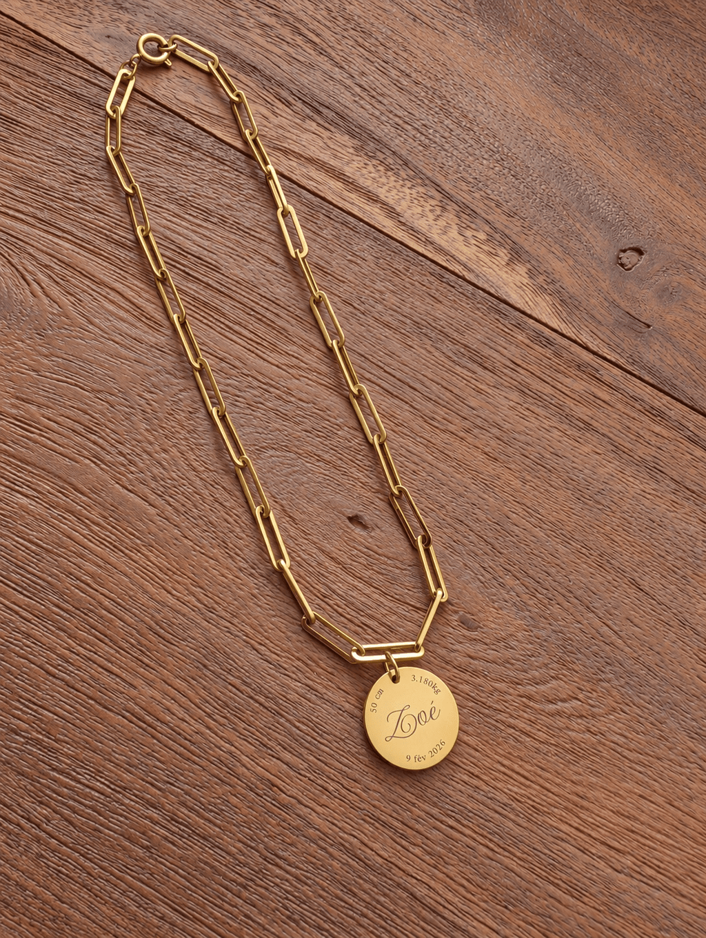 Collier de naissance complet en maille trombone dorée avec son médaillon gravé, posé sur une table en bois
