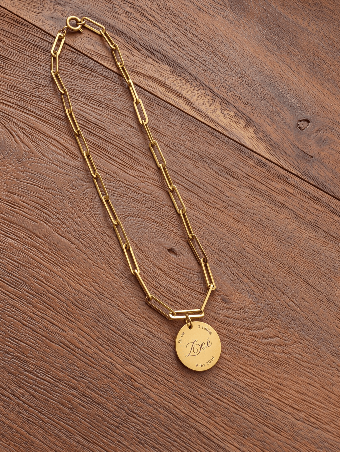 Collier de naissance complet en maille trombone dorée avec son médaillon gravé, posé sur une table en bois