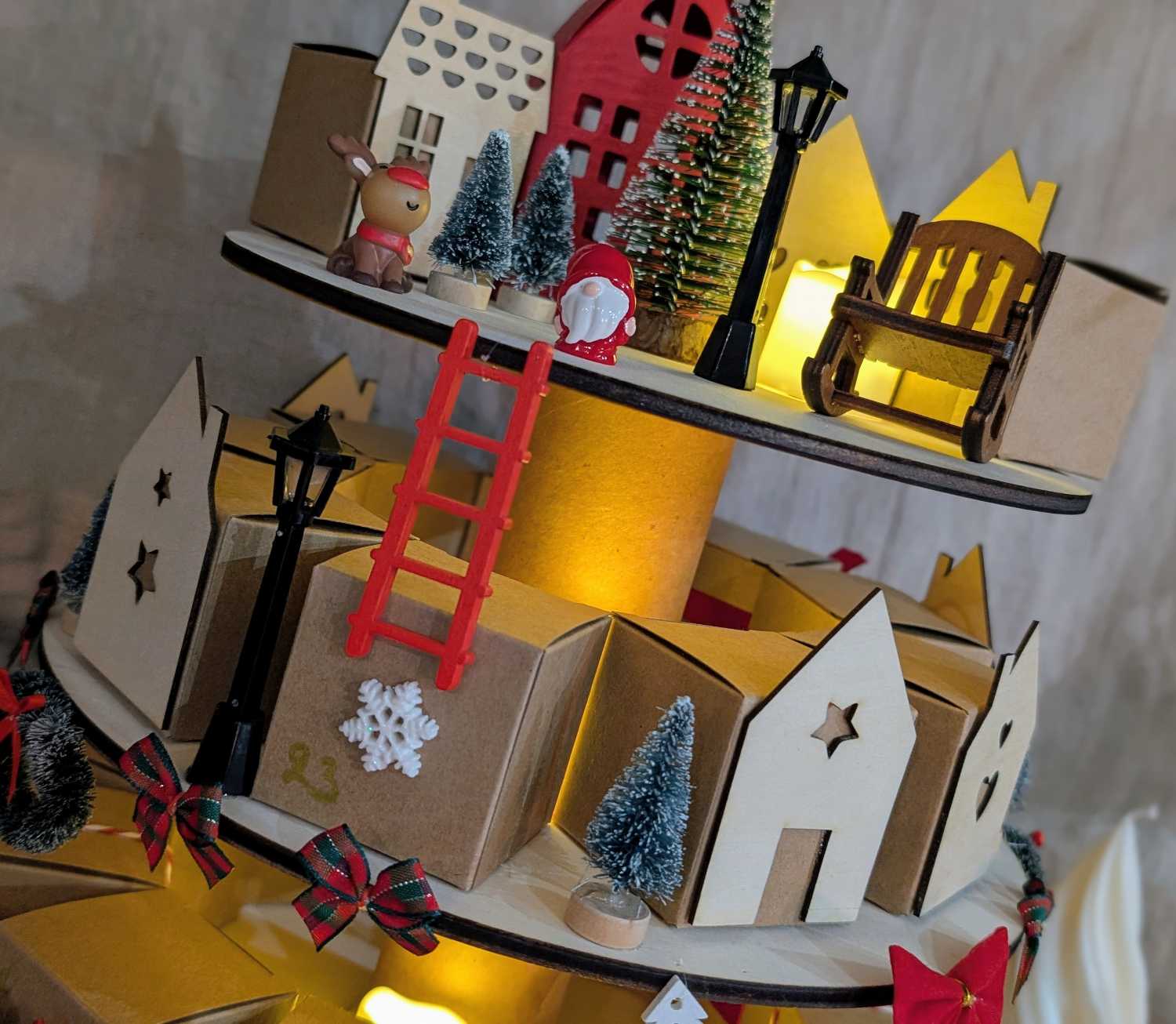 Détail décorations calendrier de l'avent : figurine renne kawaii et Père Noël rouge, mini sapin enneigé, lampadaire noir miniature et maisonnettes illuminées en arrière-plan