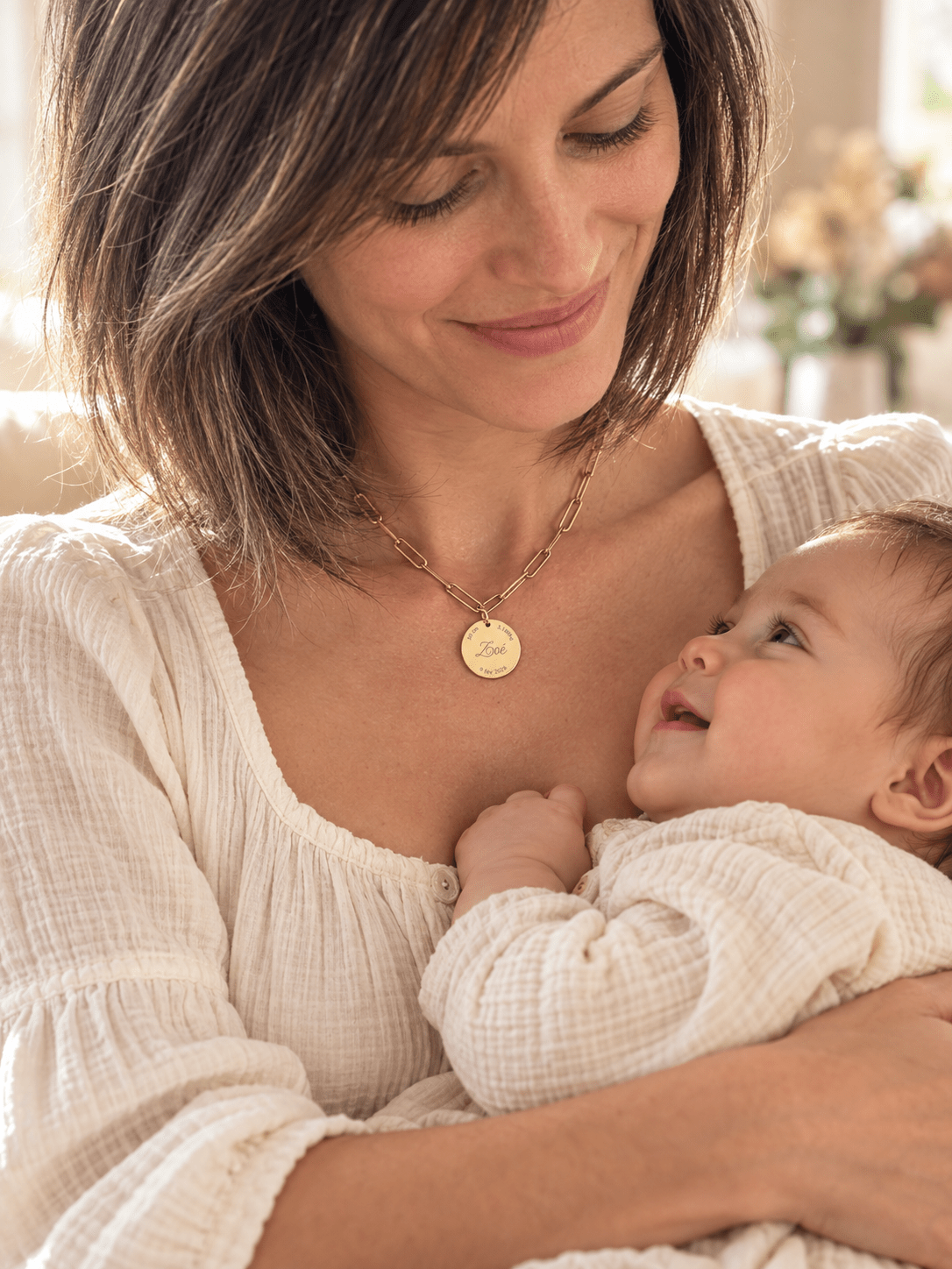 Jeune maman portant un collier de naissance personnalisé doré, médaillon gravé du prénom Zoé, son bébé dans les bras