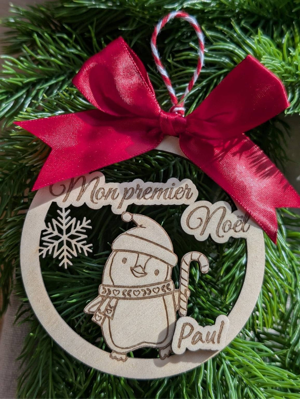 boule de Noël en bois personnalisée. Mon premier Noël. Modèle avec pingouin, flocon et prénom surmonté d'un joli nœud en satin rouge