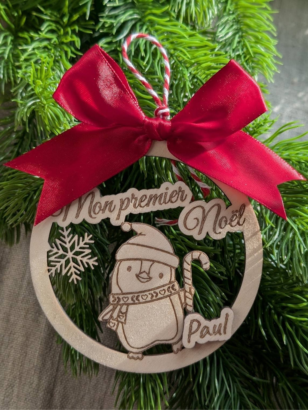 boule de Noël en bois personnalisée. Mon premier Noël. Modèle avec pingouin, flocon et prénom surmonté d'un joli nœud en satin rouge