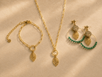 Parure bijoux dorés motif feuille : bracelet chaîne, collier long et boucles d'oreilles créoles avec perles vertes et médaillon pailleté, fond beige ensoleillé